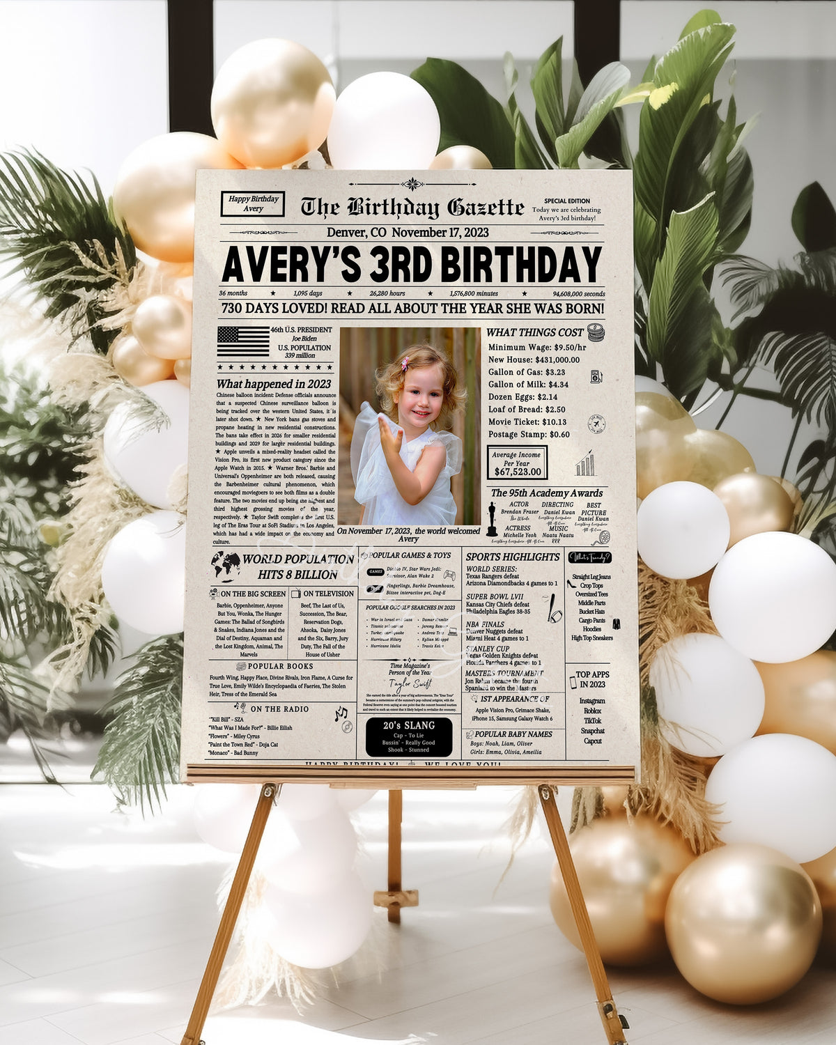 A personalized newspaper-style birthday sign on an easel, reading 'Avery's 3rd Birthday' with a photo of a young girl, displayed in a decorated event setting with white and gold balloons and greenery.
